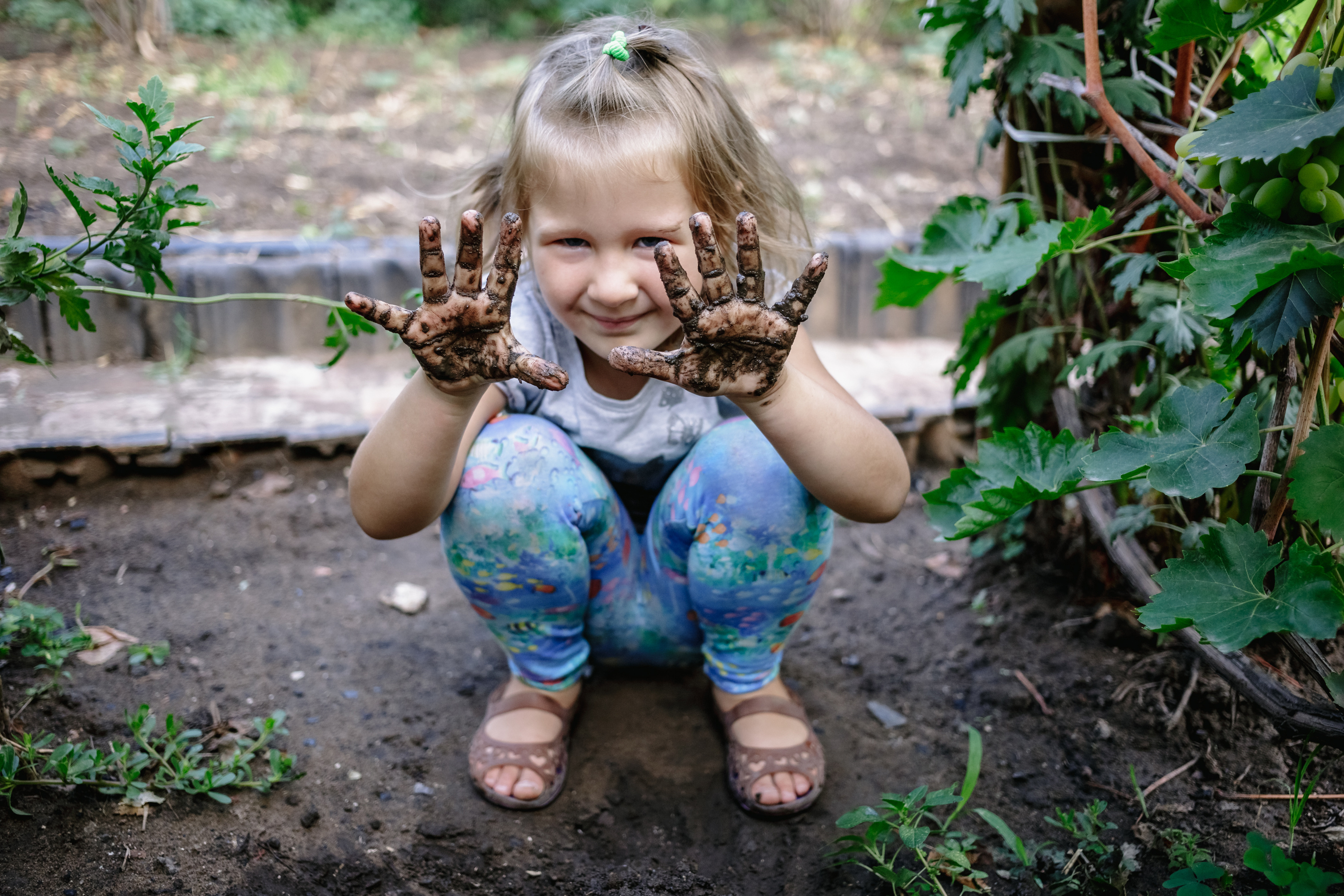 A child squats in dirt and holds up muddy hands