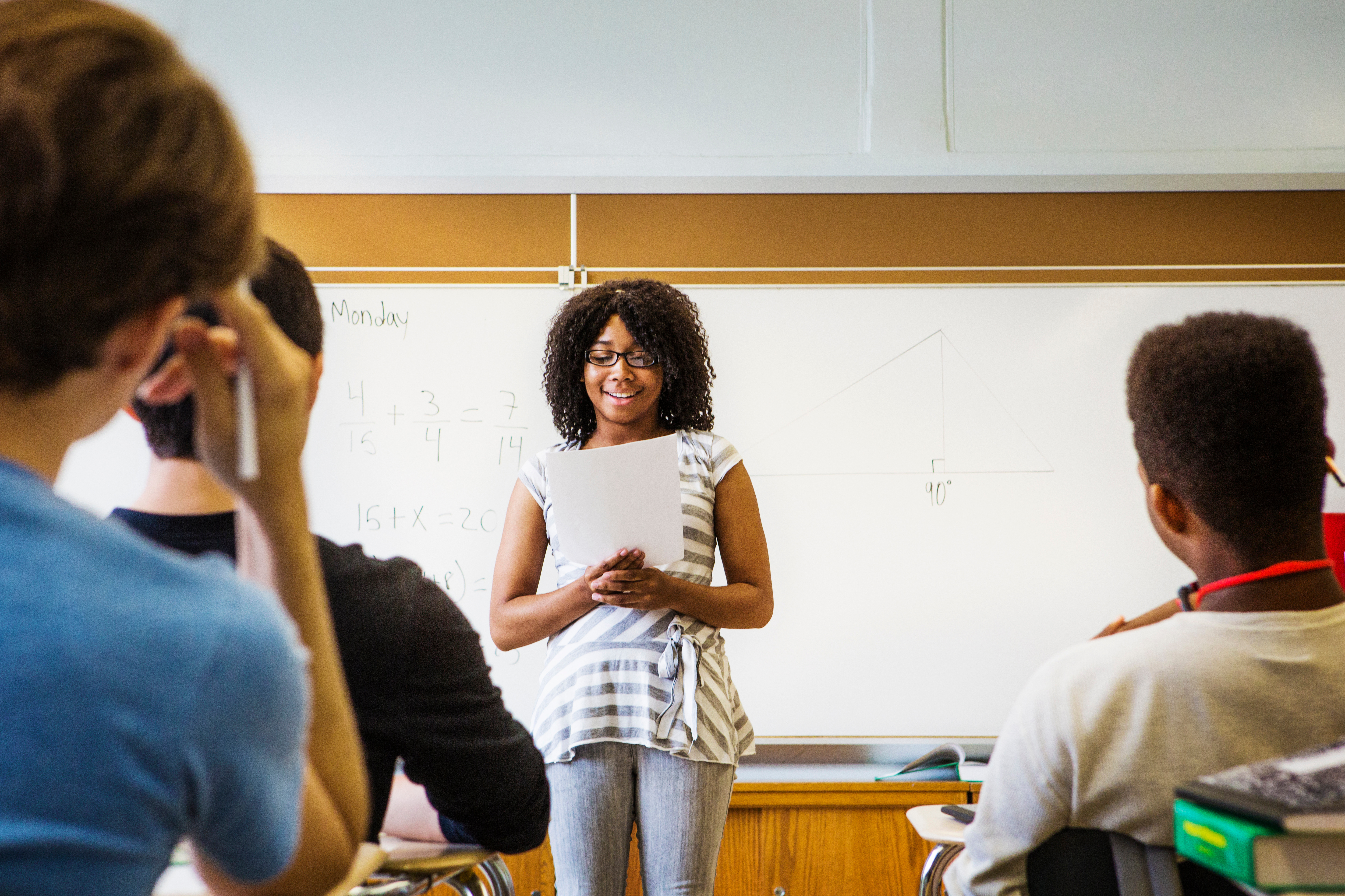 A young person stands with a paper at the front of a classroom