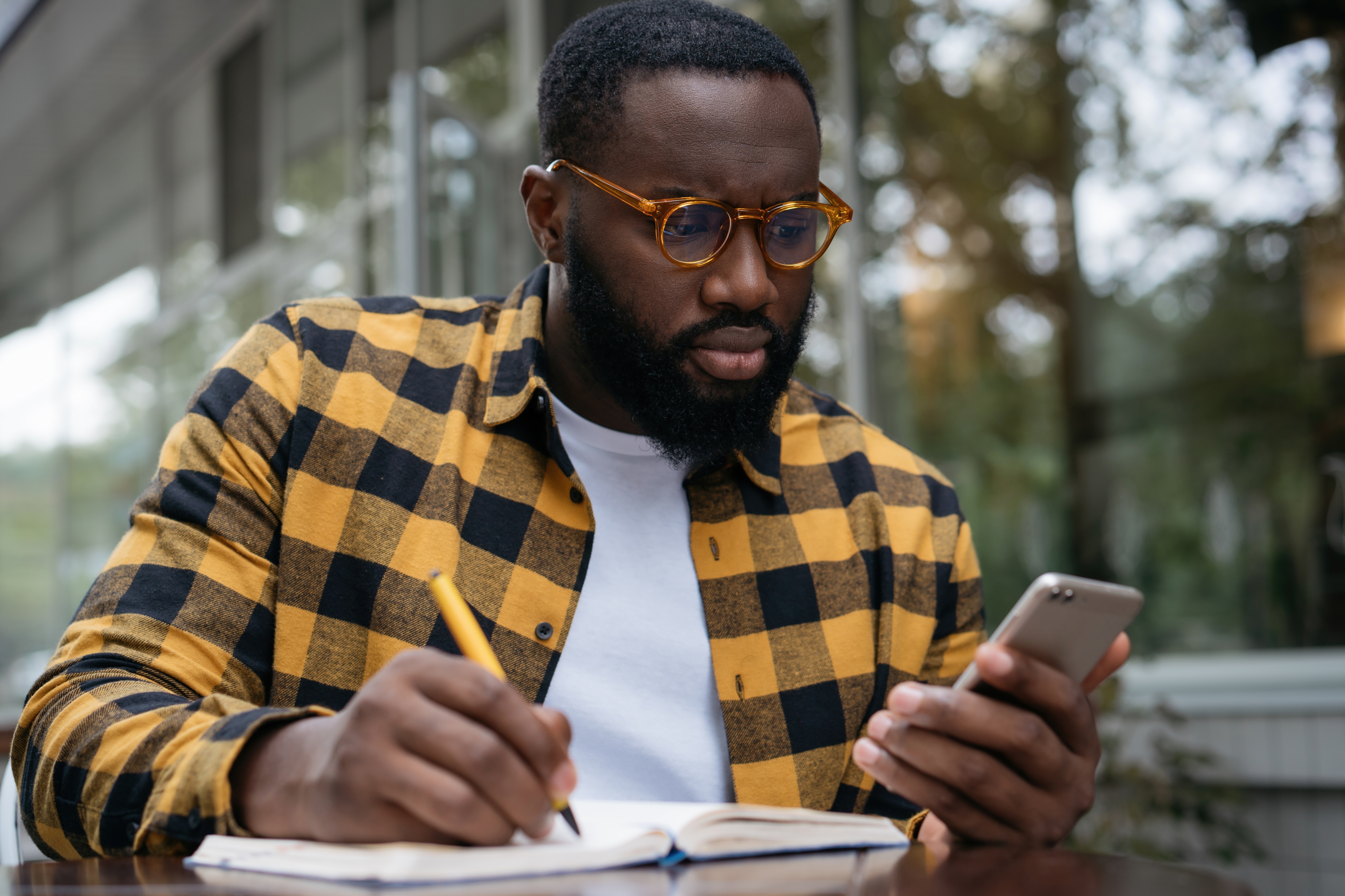 An adult sits at a table writing in a notebook while looking at a phone