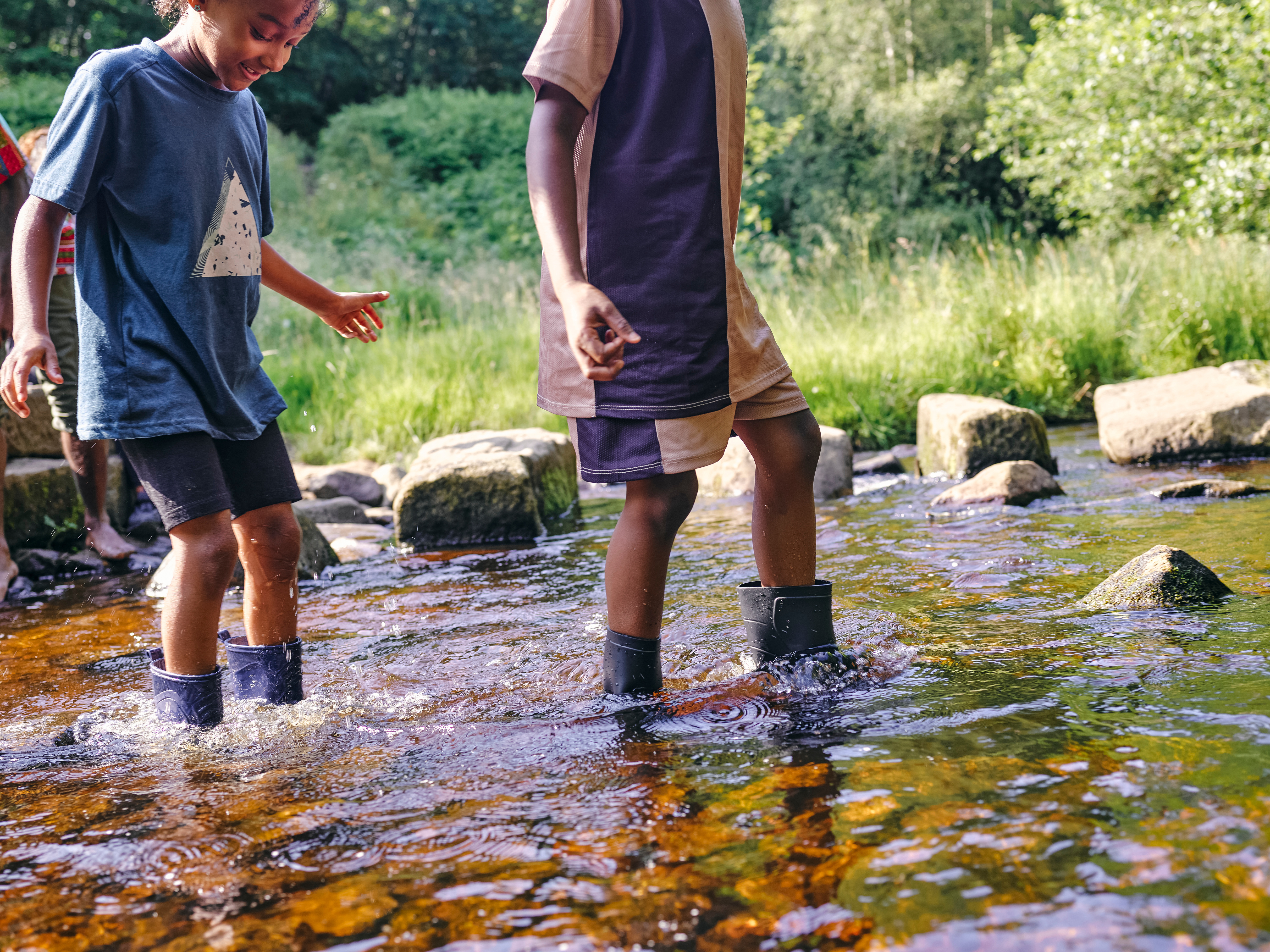 Two kids walk through a creek in rubber boots