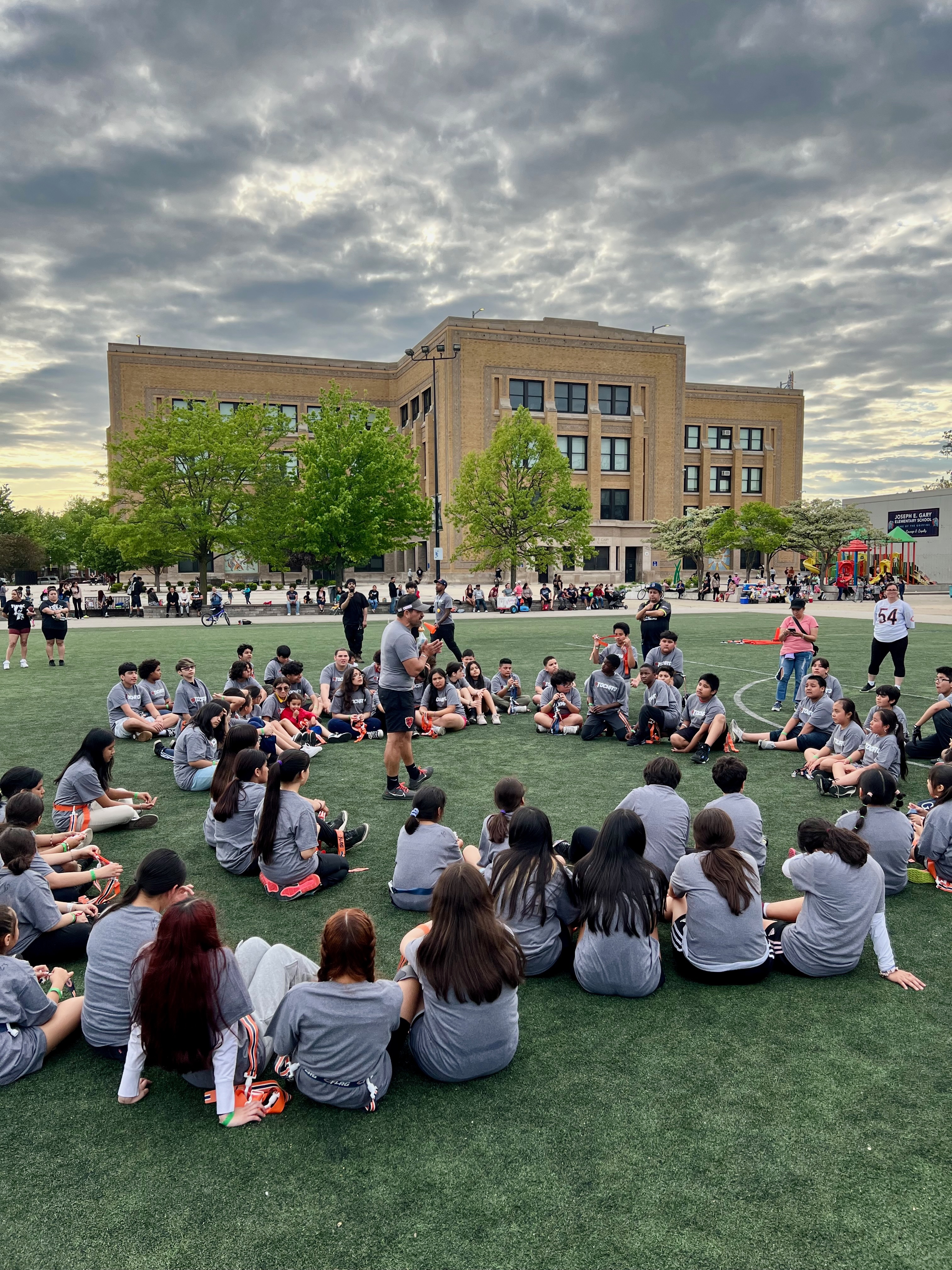 A large group of youth sit on an outdoor field looking at an adult